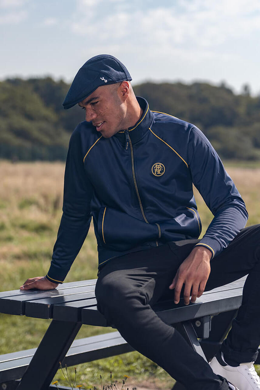 A male model sitting on a picnic bench, wearing a blue tottenham hotspur tracksuit top and flat cap