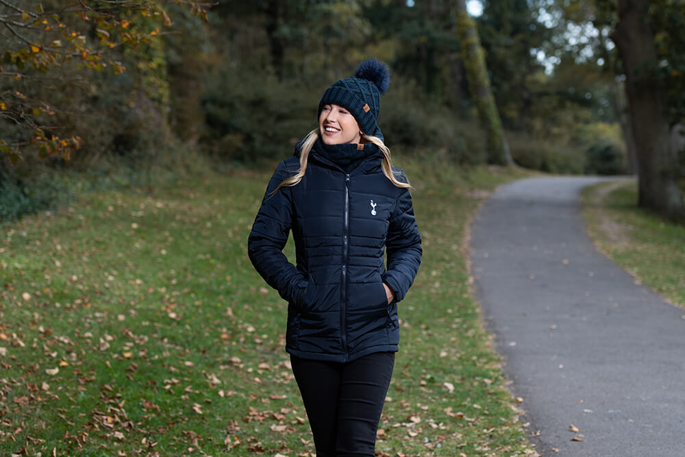 A female model walking through a park wearing a blue tottenham hotspur puffer jacket and bobble hat