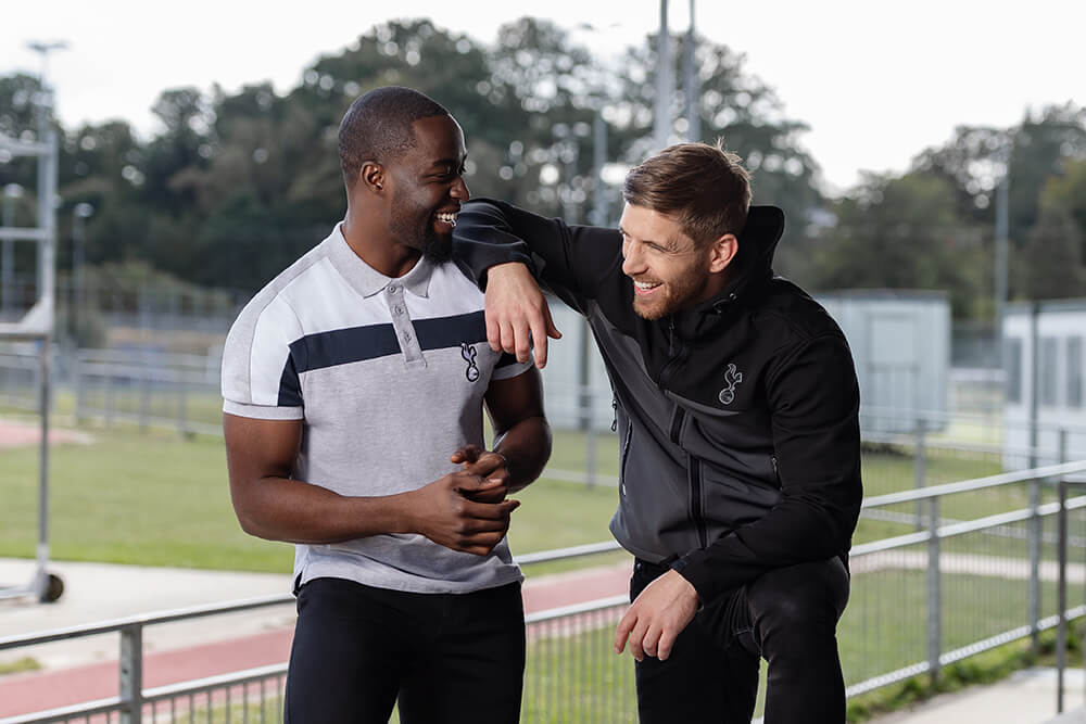 two male models standing next to a running track one is wearing a grey and black tottenham hotspur polo shirt and the other wearing a black and grey jacket